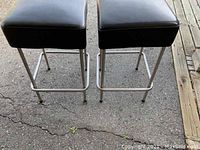 Pair of two square vinyl top stools with visible black vinyl seats and stainless steel legs on cracked pavement surface.