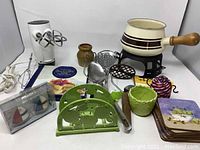 Photo showing white ceramic fondue bowl without lid on black metal stand, assorted coasters, Sunbeam electric mixer with beaters, green enamel coated napkin holder, small brown vase, kitchen utensils, colorful spiral wire trivet, and star-shaped pancake mold