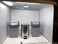 Three small trash cans displayed side by side: two gray plastic bins with white lids and one stainless steel foot pedal bin in the middle.