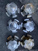 Top view of six vintage Chevy plastic chrome centre caps arranged on a textured surface, showing bowtie emblems and overall condition with visible dirt and wear.
