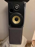 Pair of Sony speakers stacked vertically on a floor with one showing the speaker drivers and the other with the fabric grill attached.