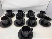 Nine black cups paired with nine matching black saucers placed on a white surface. Saucer has triangular accent visible.