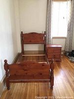 Full view of wood single bed frame with carved headboard and footboard, turned posts, wood rails, placed on hardwood floor near window with curtains.