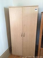 Closed pressboard cabinet against a wall with two doors and metal handles. Light wood finish with some visible wear.