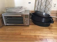 The entire kitchen lot on wooden floor, showing Emeril Lagasse toaster oven and three black pans stacked together.