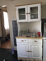 Front view of white kitchen hutch with glass doors on top, wooden top, lower cabinets, and drawer. Several glass items on counter.
