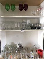 Full view of the bar glassware, mugs, shakers, and accessories arranged on white shelves showing the variation in glass styles and colors including green and dark red wine glasses, clear stemware, and a red mug.