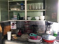 Photo of kitchen cabinet interior showing glasses, bowls, plates, appliances, and utensils on counter and shelves.