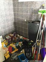 Wide shot of corner floor area with cleaning tools including mop handles, a rolled heavy duty mat, plastic bucket, and assortment of cleaning products spread out on the floor.