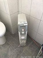 Front corner view of white oil-filled heater on castors in a tiled bathroom corner near a toilet.