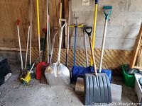 Front view of assortment of garden tools and shovels arranged against wall indoors on concrete floor.
