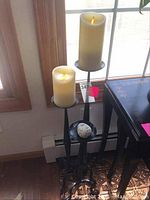 Photo showing three black metal candlesticks on the floor near a window, two with cream-colored flameless battery candles lit, one with decorative stone sphere.