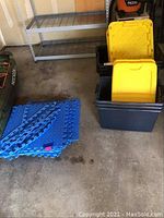 Storage tubs stacked with yellow lids inside a garage next to a stack of blue interlocking floor mats with edges.