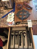 Drawer with 20-piece oiled bronze flatware set including forks, spoons, and knives. Also visible wooden cutting board, ceramic coasters, colorful wooden kitchen tools, and metal trivet on countertop.