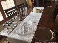 Full view of dining table with glass jars, Christmas tree figurines, oil lamp, vase, and clear glass serving trays along a white table runner.