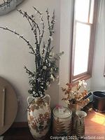 Large ceramic vase with faux flower branches and surrounding smaller ceramic pieces photographed in front of a window and wall clock.