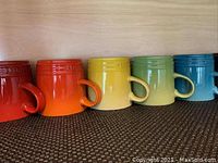 Six Le Creuset mugs in multiple bright colors lined up on a shelf.