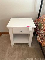 Front angle view of one white laminate bedside table next to bed, showing drawer and shelf