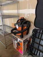 Rigid shop vac in orange and black, positioned on top of original product box with metal shelving nearby