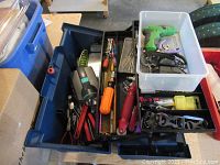 View of the open blue plastic case containing various tools including a cordless drill, pliers, screwdrivers, and assorted hand tools.