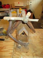 Front view showing the birdhouse facade with entrance hole, small triangular roof overhang, and natural wood branch perch in front.