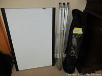 White magnetic whiteboard leaning against wall, aluminum collapsible tripod stand, and black carrying bag with markers visible inside.