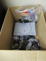 Wide view of the box showing the PlayStation console, four controllers, cables, and a plastic bag with games and manuals.