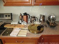 Wide view of assorted kitchenware on countertop including brass plates, mugs, beverage carafes, serving utensils, and boxes of small flatware and spoons.