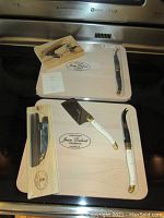 Overview of multiple Jean Dubost wooden cheese boards and Laguiole knives, some in packaging, arranged on a kitchen counter.