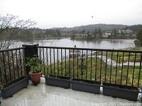 Photo of outdoor balcony corner with four black rectangular plastic garden planters placed in a row along metal railing overlooking wooded area and water body.