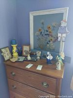 Photo of dresser top with five angel figurines, framed knitted flower art with doll, large framed flower artwork, yellow candle holder with blue candle, and decorative cat board.