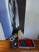 Linens on hanger and folded in plastic tote and cardboard box on floor, plus knit blanket, all near closet floor