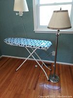 Metal legged ironing board with blue and white floral cover standing next to a metal based floor lamp with cream-colored lampshade.