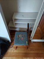 View of three wooden shoe racks stacked and placed near a wall, plaster wall showing minor marks. Blue upholstered foot stool with floral embroidery is in front.