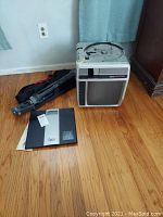 Photo showing Telex slide projector, black Epica scale, and folded Slik tripod on wooden floor near wall and curtains.