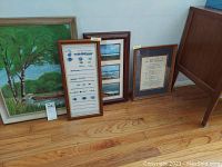 Four framed wall art pieces shown on a hardwood floor: a large landscape painting with green trees, a knitting pattern in a vertical frame, a boat scene in horizontal frame, and a framed poem with blue and gold matting.