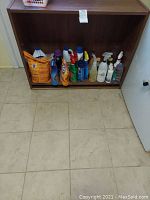 Assorted cleaning liquids and sprays placed under a wooden shelf on the floor