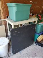Overall view of the lot showing a large green Sterlite plastic storage tub on top of a beige three-tier plastic shelving unit. Black folding card table leaning against the shelving unit.