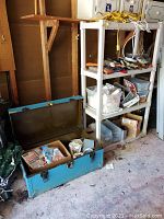Photo showing shelving unit filled with tools, plastic boxes, cords on top, and blue metal foot locker open with miscellaneous contents