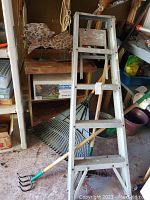 Photo showing metal 5 ft step ladder leaning against wooden rolling cart with floral cushion, large garden rake, hand cultivator tool, and part of the soaker system box