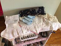 Overview of linens including a floral afghan, various doilies, and an eyelet tablecloth spread out with napkins and rings on top.