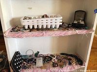 Wide view of white shelf containing multiple pieces of jewelry including pinned display trays and scattered necklaces and bracelets on pink cloth and shelves.