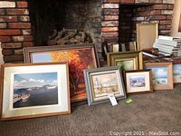 Seven framed wall art pieces displayed in front of a brick fireplace on a carpeted floor. Frames of varied styles and sizes are shown, some stacked behind others.