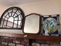 Three items on a fireplace mantel: a black framed arched mirror, a brown wooden framed mirror, and a stained glass panel with floral pattern.