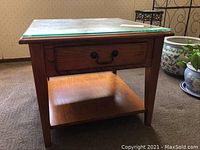 Front view of wooden end table with glass top placed on carpeted floor. Shows drawer, leg style, and bottom shelf.