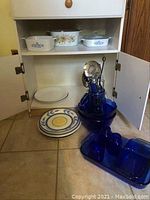 Open kitchen cabinet with 3 CorningWare casserole dishes on shelves, 7 assorted plates on floor, and blue glass items including bowls and a divided tray on floor.