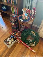 Wide view showing stacked holiday gift boxes, artificial wreath, assorted ornaments on floor, and small wooden accent table by the window.