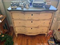 Front view of the three drawer dresser showing distressed off-white crackle finish, missing knobs on top drawer, curved drawer fronts, and stone-look dark top.