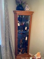 Full front view of the tall corner oak curio cabinet with glass door and decorative groove beveled etched glass front pane, showing contents inside and wood grain finish.