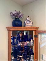 Full view of curio cabinet shelf with multiple blue glass vases, decanters, stemware and Kutani ceramic jar on top shelf and inside cabinet.